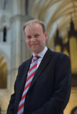 A Man standing lookng down at the camera with an organ in the background behind him